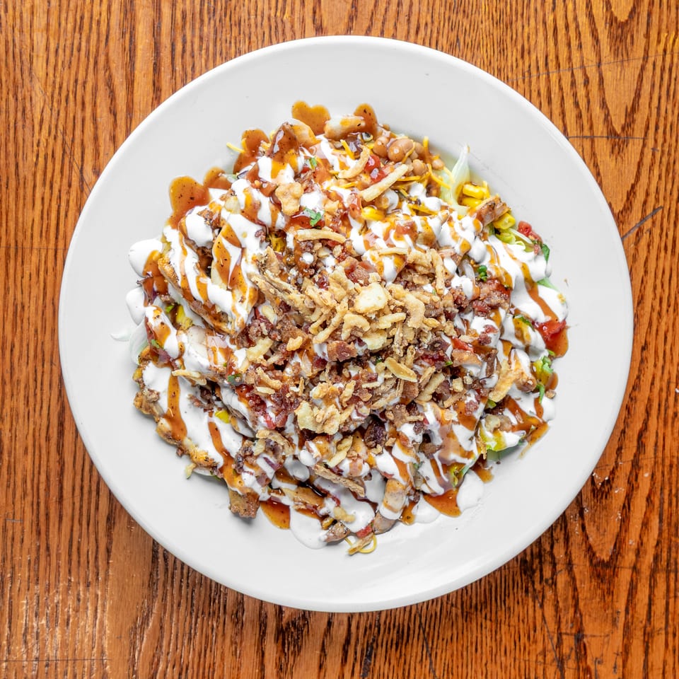 Loaded plate of fries topped with grilled meat, beans, corn, lettuce, crispy onions, bacon, cheese, and creamy sauces.