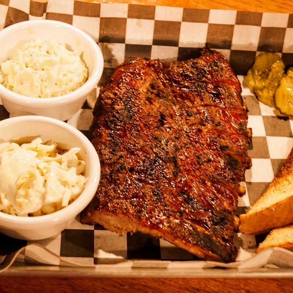 A metal tray with checkered paper holds a rack of ribs, two bowls of coleslaw, pickles, and toast.