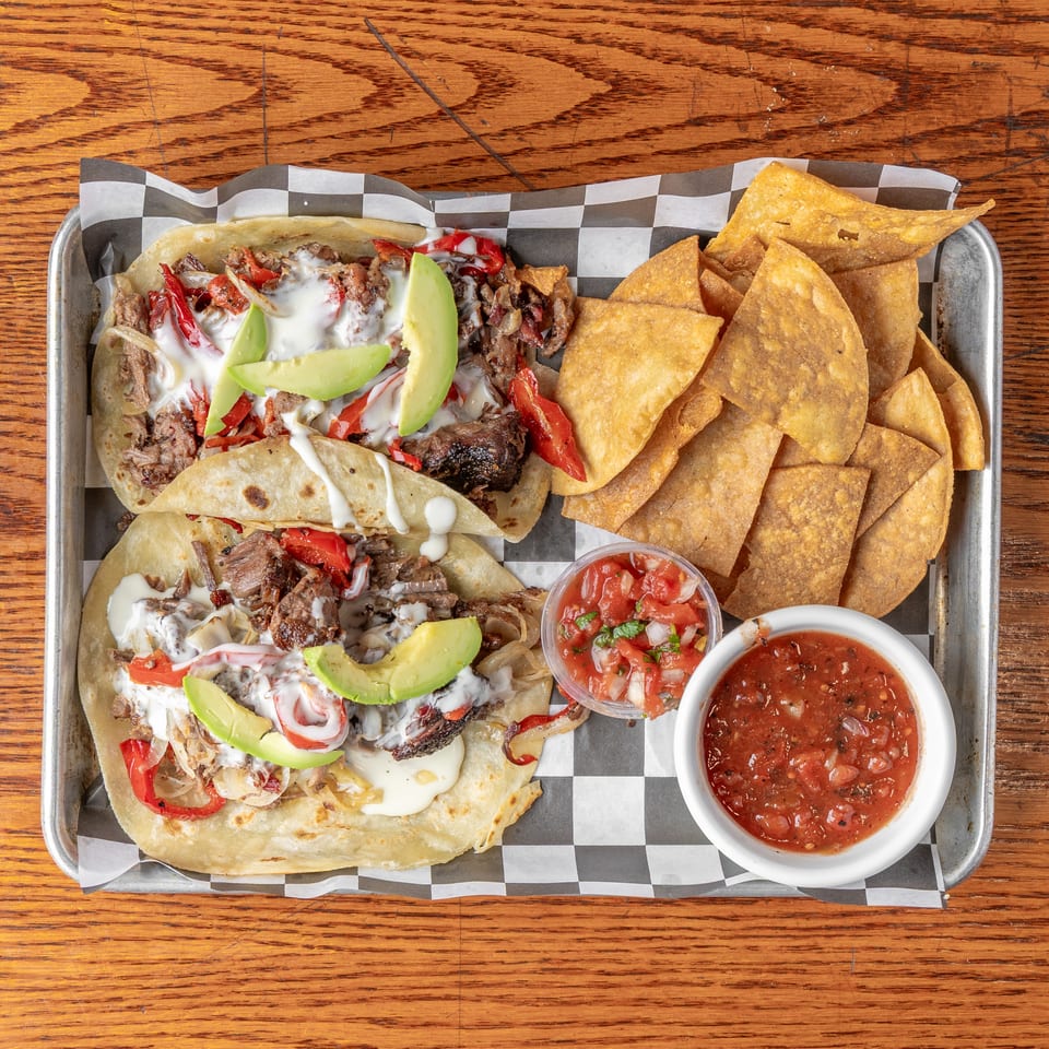 Overhead view of steak tacos topped with avocado and cream, served with tortilla chips, pico de gallo, and salsa.