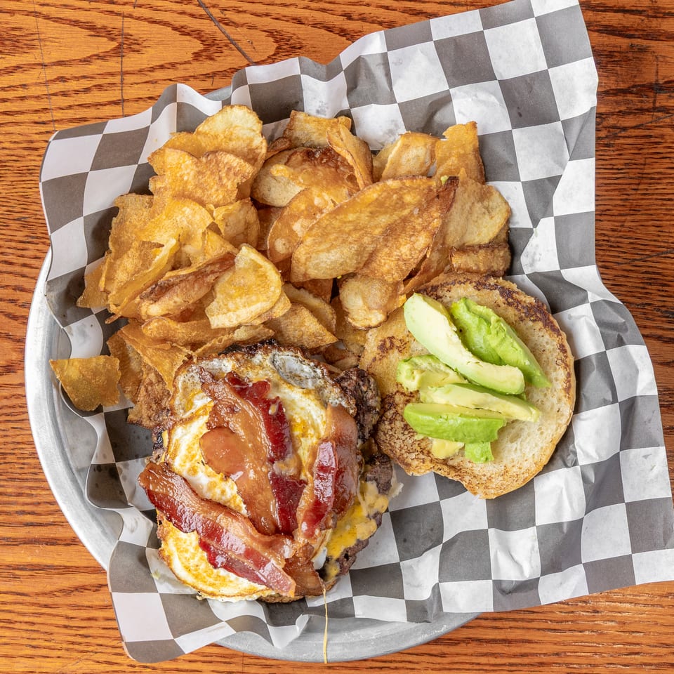 Overhead view of a bacon cheeseburger with avocado and crispy potato chips served in a checkered-lined basket.