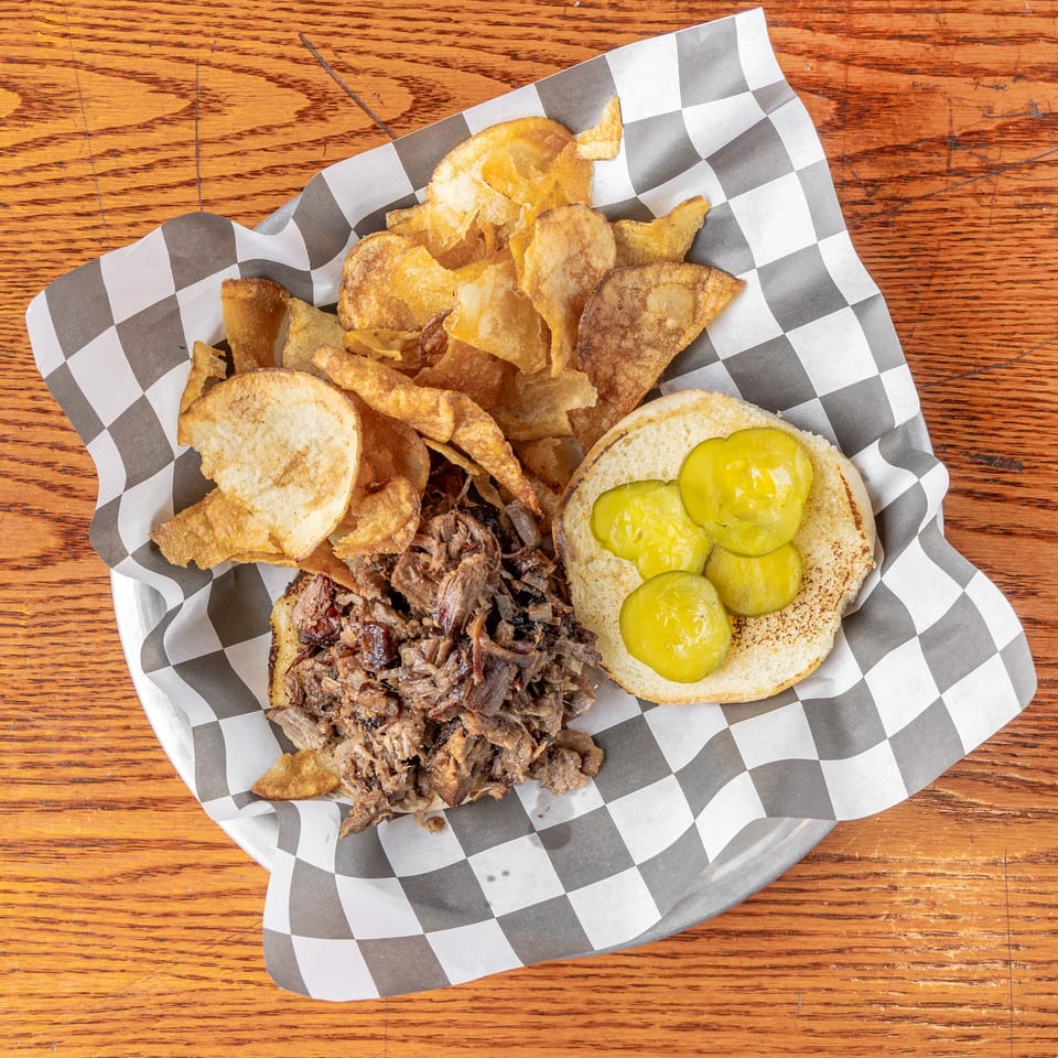 Pulled beef sandwich with pickles and crispy potato chips served in a checkered basket on a wooden table.