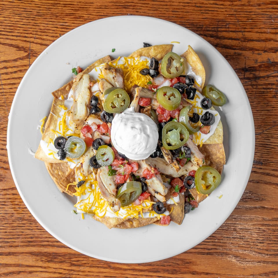 Loaded chicken nachos topped with melted cheese, sour cream, jalapeños, black olives, and pico de gallo on a white plate.