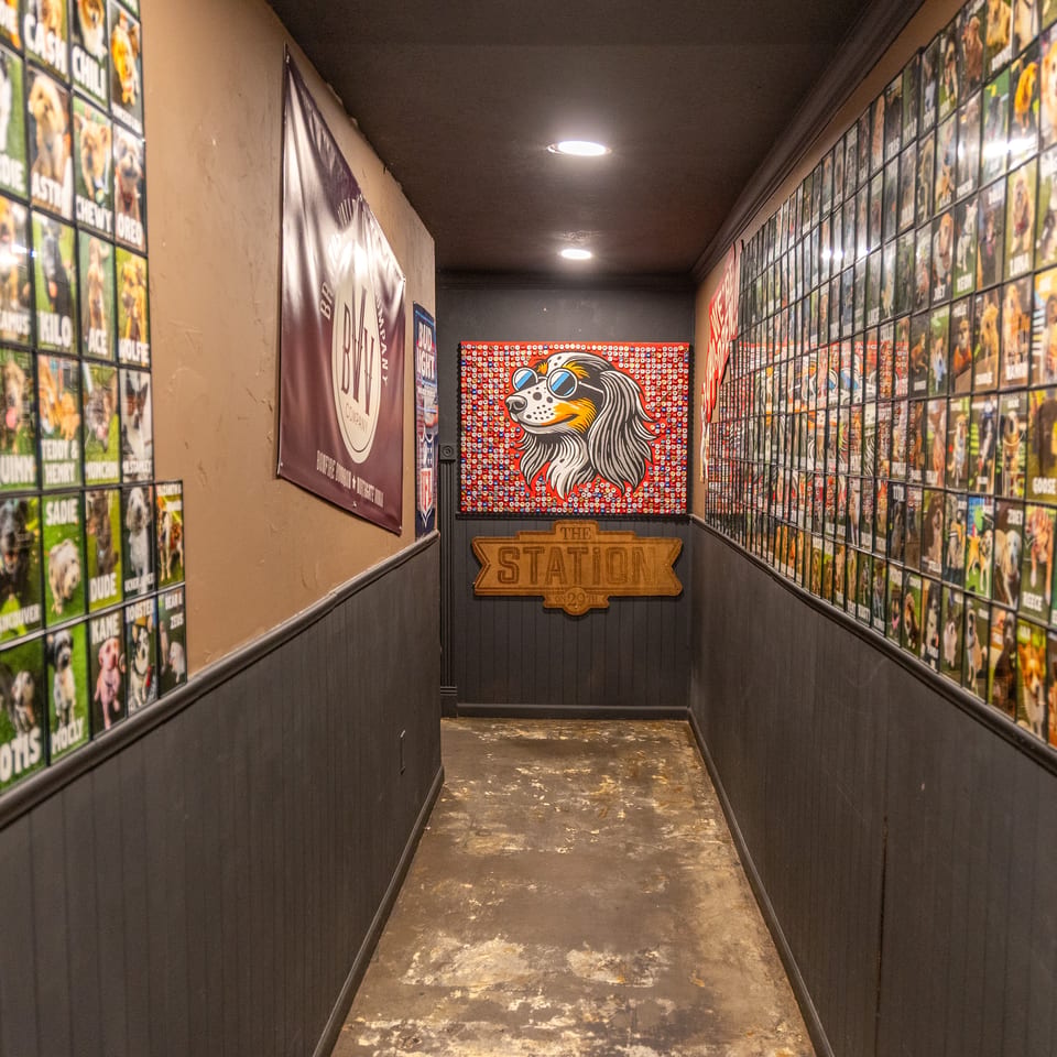 Narrow restaurant hallway covered with dog portraits and names, leading to a colorful sunglasses-wearing dog mural and Station sign.