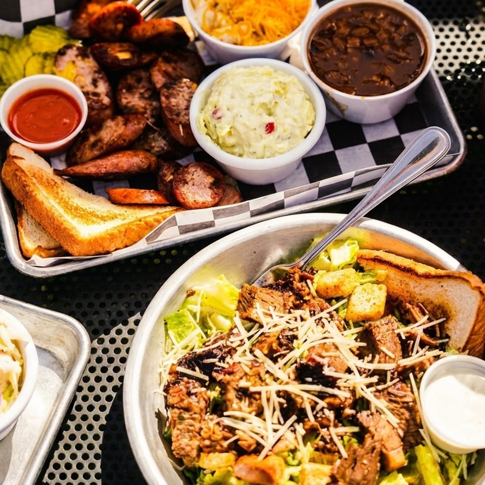 A BBQ platter with sausage, sides, and toast, alongside a brisket salad and drinks.