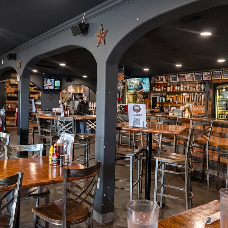 Rustic restaurant interior with wooden tables, metal chairs, arched columns, bar taps, liquor shelves, and televisions.
