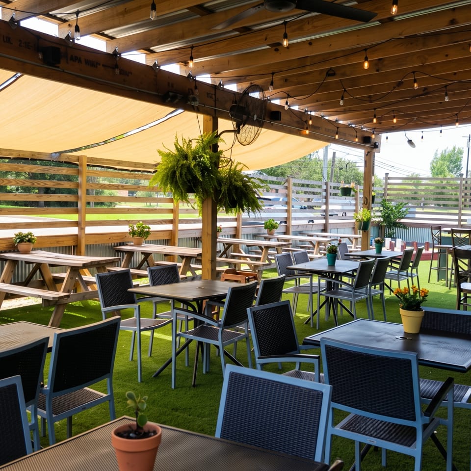 Spacious outdoor restaurant patio with picnic tables, modern seating, potted plants, string lights, and wooden sunshade.