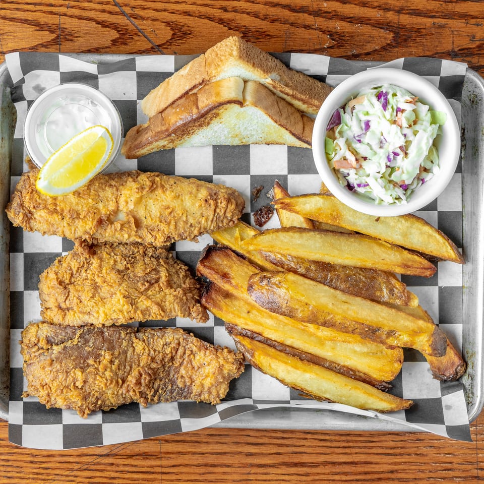 Tray of crispy fried fish with potato wedges, coleslaw, tartar sauce, lemon, and toasted bread on checkered paper.