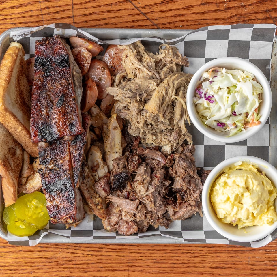 Overhead view of a barbecue platter with ribs, sausages, pulled pork, bread, pickles, coleslaw, and potato salad.