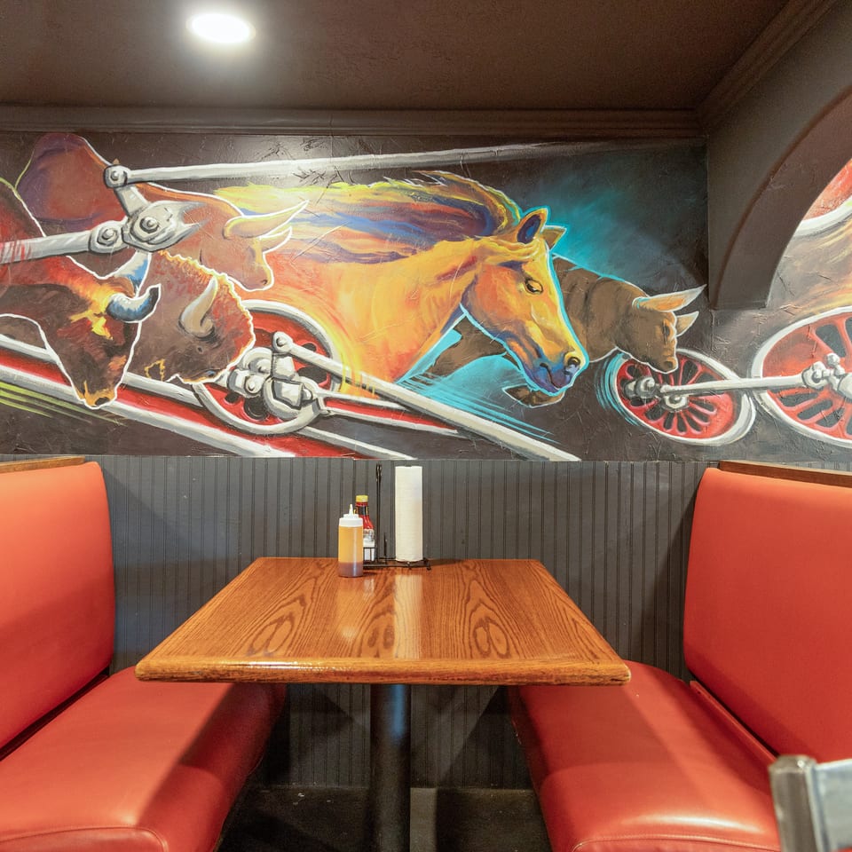 Restaurant booth with red vinyl seating, wooden table, condiments, and colorful mural of racing farm animals.